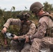 Two military personnel in desert camouflage uniforms are crouched in an outdoor field environment, examining what appears to be a drone controller or remote control device. Both individuals are wearing tactical gear including helmets, body armor, and equipment vests. The setting appears to be a training exercise or field operation in a scrubland environment.