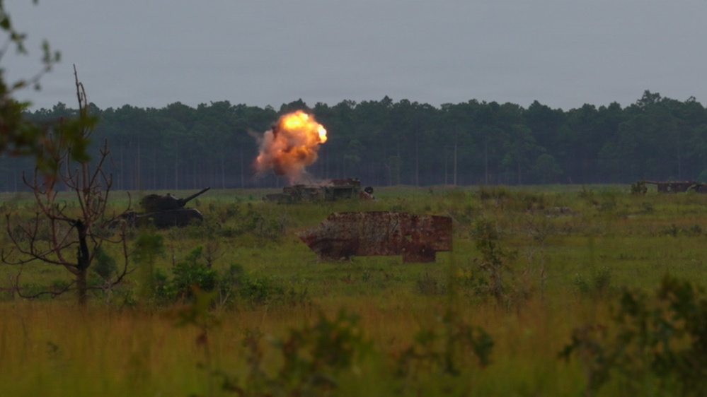 The image shows a military training exercise on an open field with armored vehicles and an active explosion or weapon discharge visible in the mid-ground. A large orange fireball is visible near what appears to be a self-propelled artillery piece or armored vehicle. The scene is set against a treeline backdrop under an overcast sky, consistent with a military training range environment.