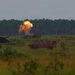 The image shows a military training exercise on an open field with armored vehicles and an active explosion or weapon discharge visible in the mid-ground. A large orange fireball is visible near what appears to be a self-propelled artillery piece or armored vehicle. The scene is set against a treeline backdrop under an overcast sky, consistent with a military training range environment.