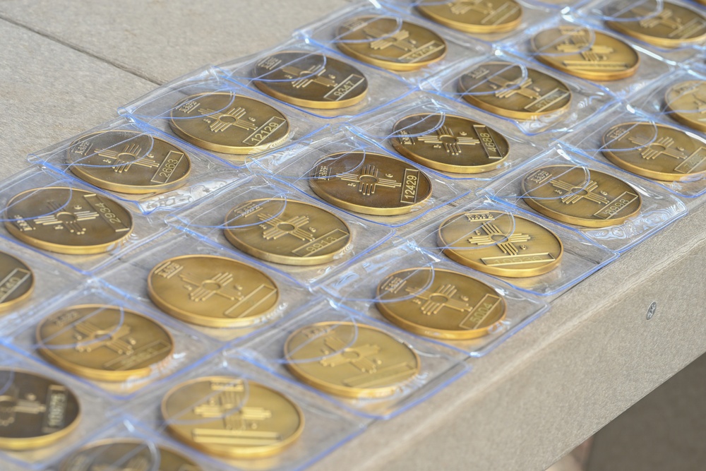 The image shows a large collection of gold-colored medallions or challenge coins arranged in individual clear plastic packaging on a light-colored table. Each coin features an identical embossed design that appears to include a Zia sun symbol, consistent with New Mexico state imagery, along with serial numbers. The coins are laid out in a grid-like pattern, suggesting they are being prepared for distribution or display.