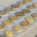 The image shows a large collection of gold-colored medallions or challenge coins arranged in individual clear plastic packaging on a light-colored table. Each coin features an identical embossed design that appears to include a Zia sun symbol, consistent with New Mexico state imagery, along with serial numbers. The coins are laid out in a grid-like pattern, suggesting they are being prepared for distribution or display.