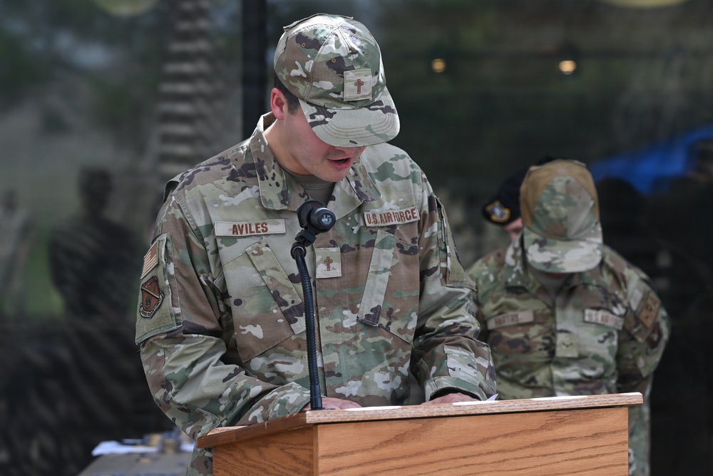 A U.S. Air Force service member named Aviles stands at a wooden podium speaking into a microphone, appearing to read from documents. He is wearing OCP (Operational Camouflage Pattern) uniform with a chaplain cross insignia visible on both his cap and uniform. A second U.S. Air Force service member is visible in the background, also in OCP uniform.