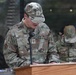 A U.S. Air Force service member named Aviles stands at a wooden podium speaking into a microphone, appearing to read from documents. He is wearing OCP (Operational Camouflage Pattern) uniform with a chaplain cross insignia visible on both his cap and uniform. A second U.S. Air Force service member is visible in the background, also in OCP uniform.