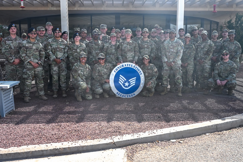 A large group of U.S. Air Force personnel in OCP (Operational Camouflage Pattern) uniforms pose for a group photograph. Three individuals in the front row are kneeling and holding a circular sign reading 'Congratulations Selectees' featuring the Staff Sergeant rank insignia. This appears to be a promotion selectee recognition photo taken outdoors at a military installation.