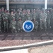 A large group of U.S. Air Force personnel in OCP (Operational Camouflage Pattern) uniforms pose for a group photograph. Three individuals in the front row are kneeling and holding a circular sign reading 'Congratulations Selectees' featuring the Staff Sergeant rank insignia. This appears to be a promotion selectee recognition photo taken outdoors at a military installation.