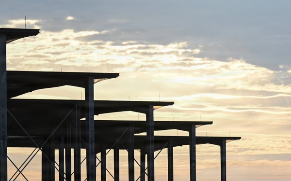 The image shows silhouettes of military personnel walking across an airfield tarmac at dawn or dusk. A row of open-sided aircraft shelters or sunshades lines the left side, with what appear to be military jet aircraft parked beneath them. The warm golden sky contrasts with the dark silhouetted figures and structures.