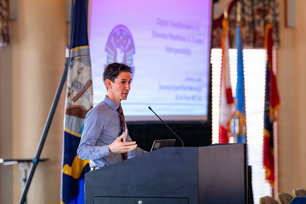 A man is delivering a presentation at a podium in what appears to be a formal or institutional setting. Behind him is a projected slide featuring an Egyptian pharaoh mask graphic with partially legible text about 'Digital Transformation' and 'Elevated Readiness &amp; Trauma Interoperability'. Multiple flags, including what appears to be a Virginia state flag, are visible in the background.