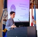 A man is delivering a presentation at a podium in what appears to be a formal or institutional setting. Behind him is a projected slide featuring an Egyptian pharaoh mask graphic with partially legible text about 'Digital Transformation' and 'Elevated Readiness &amp; Trauma Interoperability'. Multiple flags, including what appears to be a Virginia state flag, are visible in the background.