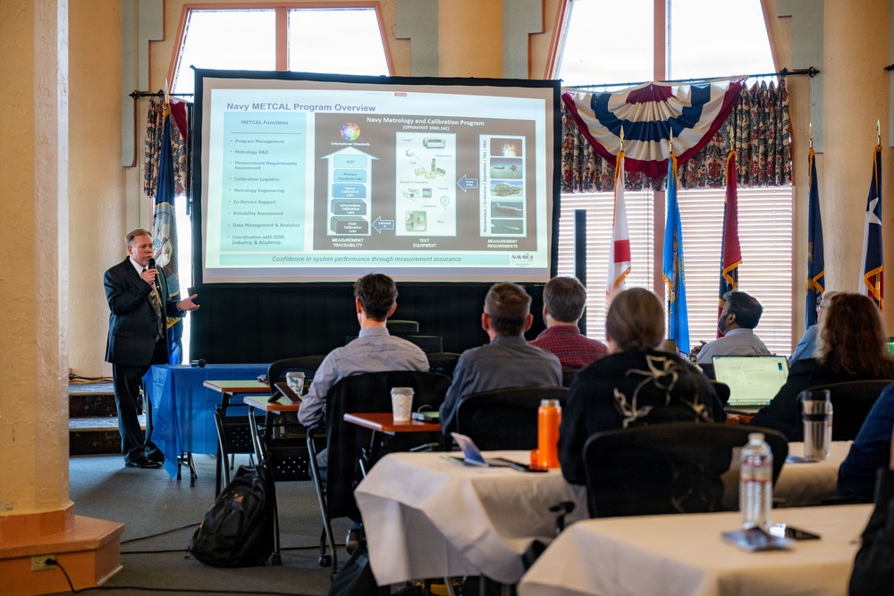 A presenter is delivering a briefing on the 'Navy METCAL Program Overview' to a small audience seated at tables in a formal meeting room. The projected slide displays information about the Navy Metrology and Calibration Program. The setting appears to be an official government or military venue, indicated by the flags and formal decor.