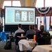 A presenter is delivering a briefing on the 'Navy METCAL Program Overview' to a small audience seated at tables in a formal meeting room. The projected slide displays information about the Navy Metrology and Calibration Program. The setting appears to be an official government or military venue, indicated by the flags and formal decor.