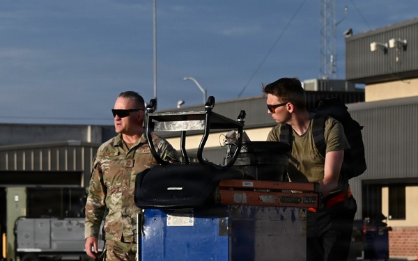 Two military personnel are moving equipment on a wheeled blue cart across what appears to be a military airfield or flight line. One individual is wearing a full camouflage uniform with tan boots, while the other is in a dark t-shirt and pants with a backpack. The background shows hangars, ground support equipment, and other personnel in the distance.