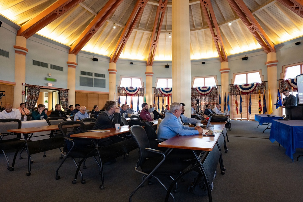 A formal meeting or conference is taking place inside a large, circular rotunda-style room with a distinctive wooden vaulted ceiling supported by decorative columns. Attendees are seated at long rectangular tables arranged in rows, facing a presenter at a podium on the right side of the room. The venue is decorated with red, white, and blue patriotic bunting and multiple flags, suggesting a government or military-affiliated event.