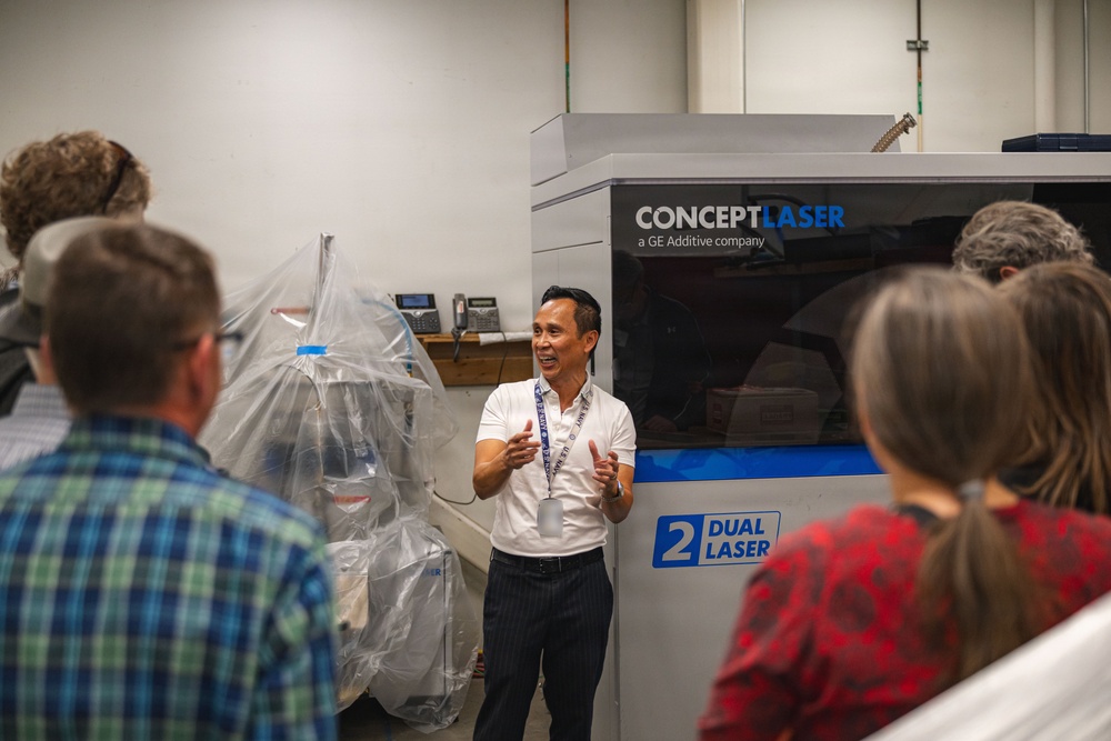 A man in a white polo shirt and lanyard is presenting or giving a tour in front of a Concept Laser (a GE Additive company) dual laser metal 3D printing machine. He is gesturing with both hands and appears to be explaining the equipment to a small group of attendees. The setting appears to be an industrial or manufacturing facility.