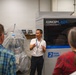 A man in a white polo shirt and lanyard is presenting or giving a tour in front of a Concept Laser (a GE Additive company) dual laser metal 3D printing machine. He is gesturing with both hands and appears to be explaining the equipment to a small group of attendees. The setting appears to be an industrial or manufacturing facility.
