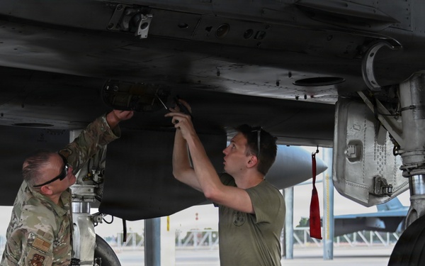 Two military personnel are working underneath a military aircraft, appearing to perform maintenance or weapons loading tasks on an underside hardpoint or munition. One individual is wearing a camouflage uniform while the other wears an olive green t-shirt with dark pants and a red safety belt. The work is taking place on a flight line, with another aircraft visible in the background.