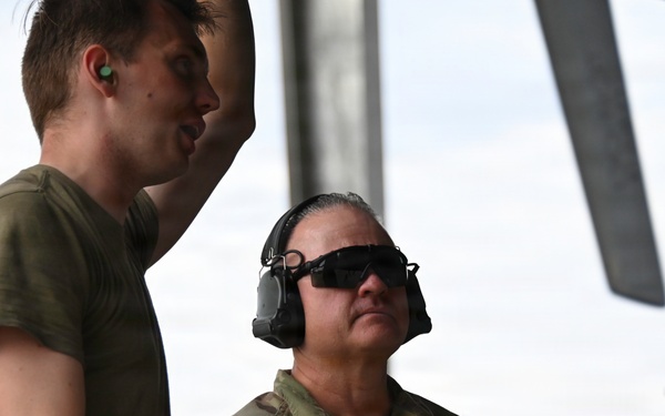 Two U.S. Air Force personnel are working near a military aircraft, likely conducting maintenance or pre-flight inspection activities. One individual wears a camouflage uniform with a name tape reading 'SCHMITS' and 'U.S. AIR FORCE' patch, along with hearing protection and sunglasses. The other person is in an olive drab t-shirt with a red safety belt and green ear plugs.