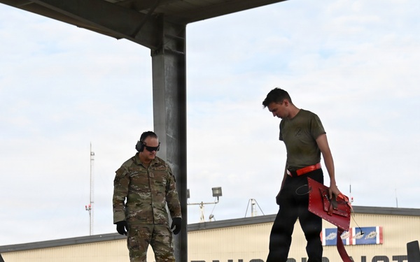 Two military personnel are working on top of a fighter jet, likely an F-16, at a California Air National Guard facility. One individual is in full camouflage uniform with hearing protection, while the other wears a olive t-shirt and carries a red tool case. The background shows a hangar with partial text reading 'CALIFORNIA AIR NAT[IONAL GUARD]'.