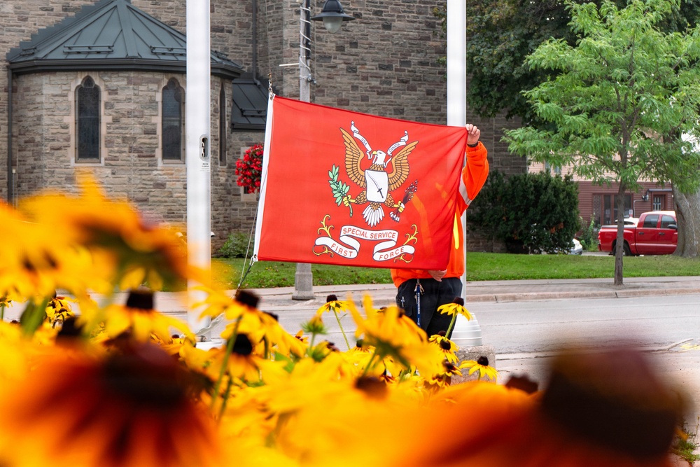 A person wearing an orange high-visibility jacket is holding up a red flag bearing the crest of the 'Special Service Force / First Force' military unit, featuring an eagle emblem. The scene is set on a street with a stone church building visible in the background, framed in the foreground by yellow and orange black-eyed Susan flowers in soft focus. The composition creates a layered depth effect between the flowers, the flag holder, and the architectural background.