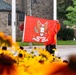 A person wearing an orange high-visibility jacket is holding up a red flag bearing the crest of the 'Special Service Force / First Force' military unit, featuring an eagle emblem. The scene is set on a street with a stone church building visible in the background, framed in the foreground by yellow and orange black-eyed Susan flowers in soft focus. The composition creates a layered depth effect between the flowers, the flag holder, and the architectural background.