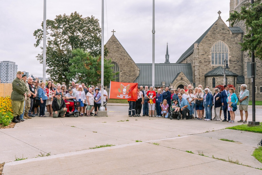 A large group of approximately 50-70 people are gathered outdoors in front of a Gothic-style stone church for what appears to be a commemorative or ceremonial event. Several individuals are holding a red flag or banner with an emblem in the center, suggesting an organizational or regimental gathering. The group includes people of various ages, some in wheelchairs or mobility devices, and at least one person in military-style attire on the far left.