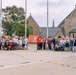 A large group of approximately 50-70 people are gathered outdoors in front of a Gothic-style stone church for what appears to be a commemorative or ceremonial event. Several individuals are holding a red flag or banner with an emblem in the center, suggesting an organizational or regimental gathering. The group includes people of various ages, some in wheelchairs or mobility devices, and at least one person in military-style attire on the far left.