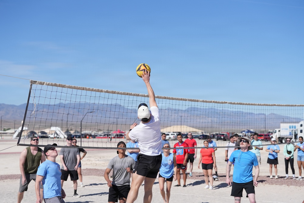 A group of people are playing outdoor volleyball on a dirt court in what appears to be a desert environment. A player in a white shirt and cap is reaching above the net to spike or block a yellow volleyball. Spectators in light blue shirts and casual attire watch from the sidelines, with mountains and vehicles visible in the background.