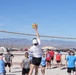 A group of people are playing outdoor volleyball on a dirt court in what appears to be a desert environment. A player in a white shirt and cap is reaching above the net to spike or block a yellow volleyball. Spectators in light blue shirts and casual attire watch from the sidelines, with mountains and vehicles visible in the background.