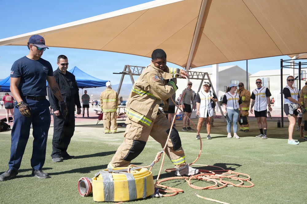 A firefighter in full turnout gear is performing a hose deployment drill on an artificial turf field at what appears to be an outdoor training event or competition. Several observers, including other firefighters and civilians, watch from the sides. The event takes place under a large shade canopy structure in what appears to be a desert environment.