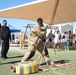 A firefighter in full turnout gear is performing a hose deployment drill on an artificial turf field at what appears to be an outdoor training event or competition. Several observers, including other firefighters and civilians, watch from the sides. The event takes place under a large shade canopy structure in what appears to be a desert environment.
