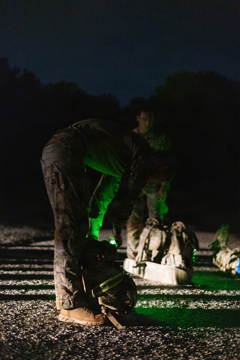 A nighttime military or tactical training exercise captured on what appears to be a gravel surface. Personnel in camouflage uniforms are bent over equipment, illuminated by green chemlight/glow stick light sources. Multiple rucksacks and tactical gear are visible on the ground.