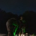 A nighttime military or tactical training exercise captured on what appears to be a gravel surface. Personnel in camouflage uniforms are bent over equipment, illuminated by green chemlight/glow stick light sources. Multiple rucksacks and tactical gear are visible on the ground.