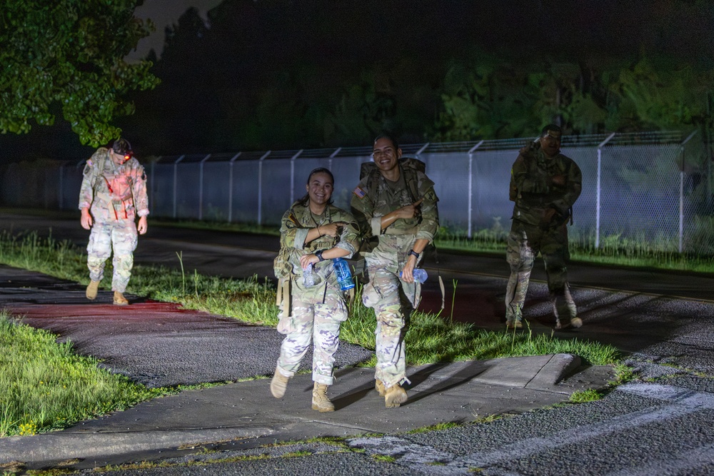 Four military personnel in camouflage uniforms are walking along a paved path at night, illuminated by artificial lighting. Two soldiers in the foreground are smiling and appear to be in good spirits, carrying water bottles, while two others follow behind. The setting appears to be a military installation or base, identifiable by the security fencing in the background.