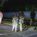 Four military personnel in camouflage uniforms are walking along a paved path at night, illuminated by artificial lighting. Two soldiers in the foreground are smiling and appear to be in good spirits, carrying water bottles, while two others follow behind. The setting appears to be a military installation or base, identifiable by the security fencing in the background.