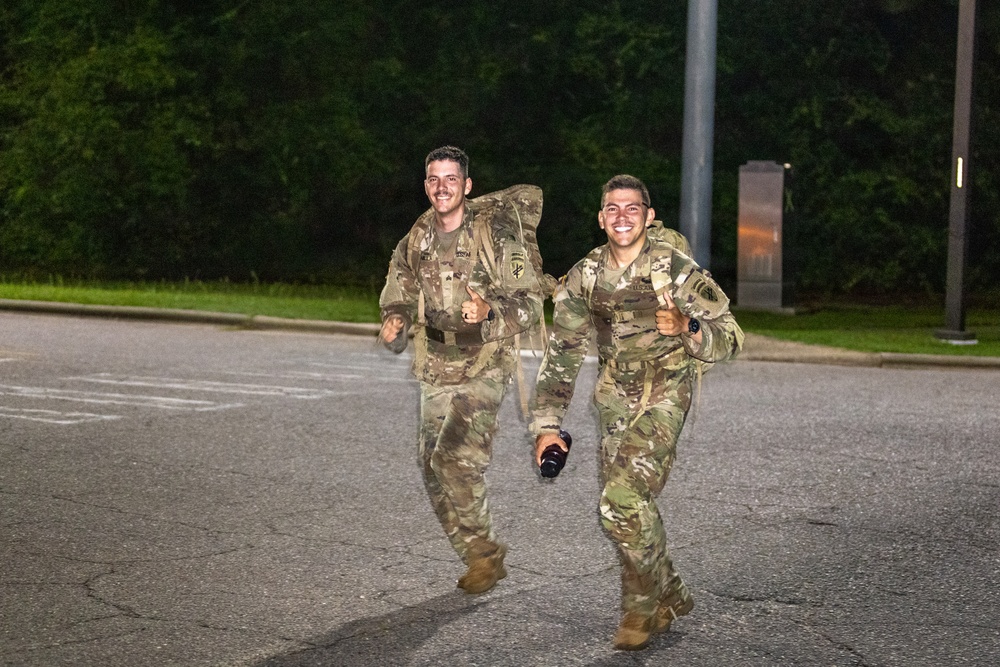 Two male soldiers in U.S. Army multicam (OCP) uniforms are running together on a paved road at night or in low-light conditions, both smiling and appearing in good spirits. They are wearing rucksacks and tactical gear, suggesting participation in a ruck march or physical training event. The setting appears to be a military installation based on the surrounding infrastructure.