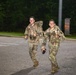 Two male soldiers in U.S. Army multicam (OCP) uniforms are running together on a paved road at night or in low-light conditions, both smiling and appearing in good spirits. They are wearing rucksacks and tactical gear, suggesting participation in a ruck march or physical training event. The setting appears to be a military installation based on the surrounding infrastructure.