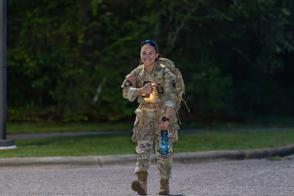 A female U.S. Army soldier in full OCP (Operational Camouflage Pattern) uniform is walking along a paved road while smiling and giving a thumbs-up gesture. She is carrying a military rucksack on her back and holding a water bottle, suggesting she is participating in a ruck march or physical training event. The setting appears to be a military installation during early morning or evening hours.