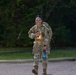 A female U.S. Army soldier in full OCP (Operational Camouflage Pattern) uniform is walking along a paved road while smiling and giving a thumbs-up gesture. She is carrying a military rucksack on her back and holding a water bottle, suggesting she is participating in a ruck march or physical training event. The setting appears to be a military installation during early morning or evening hours.