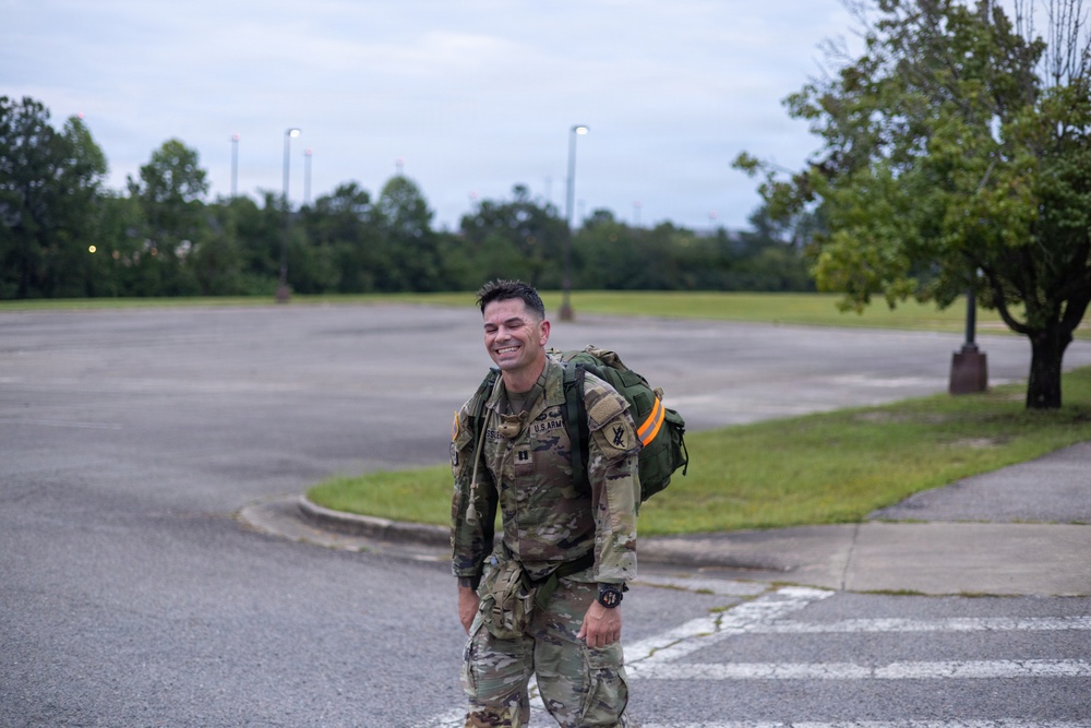 A U.S. Army soldier in full camouflage (OCP/MultiCam) uniform is walking across a paved area, smiling broadly, while carrying a large military rucksack on his back. He appears to be on a military installation, likely completing a ruck march or physical training exercise. The setting is an overcast day with a large open parking lot and green trees visible in the background.