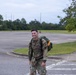 A U.S. Army soldier in full camouflage (OCP/MultiCam) uniform is walking across a paved area, smiling broadly, while carrying a large military rucksack on his back. He appears to be on a military installation, likely completing a ruck march or physical training exercise. The setting is an overcast day with a large open parking lot and green trees visible in the background.
