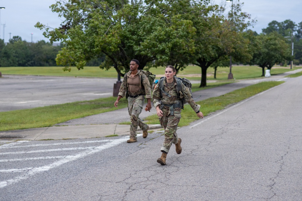 Two military personnel in camouflage uniforms are running or fast-walking along a road on what appears to be a military installation. Both are carrying large rucksacks on their backs, suggesting they are participating in a ruck march or physical training exercise. The setting includes a well-maintained grassy area with trees lining the road under an overcast sky.