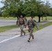 Two military personnel in camouflage uniforms are running or fast-walking along a road on what appears to be a military installation. Both are carrying large rucksacks on their backs, suggesting they are participating in a ruck march or physical training exercise. The setting includes a well-maintained grassy area with trees lining the road under an overcast sky.