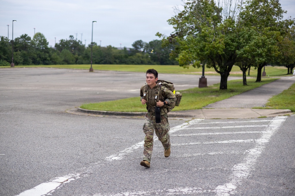 A soldier in full military camouflage (OCP/MultiCam) uniform is running across a paved road or parking lot area while carrying a large rucksack and what appears to be a rifle or similar equipment. The setting appears to be a military installation or base, identifiable by the manicured grounds, parking lots, and light poles. The soldier appears to be participating in a ruck march or physical fitness event.