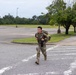 A soldier in full military camouflage (OCP/MultiCam) uniform is running across a paved road or parking lot area while carrying a large rucksack and what appears to be a rifle or similar equipment. The setting appears to be a military installation or base, identifiable by the manicured grounds, parking lots, and light poles. The soldier appears to be participating in a ruck march or physical fitness event.