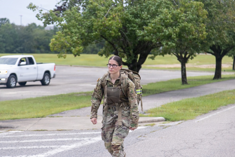 A female soldier in full military camouflage (OCP/MultiCam) uniform is walking along a paved road or parking area on what appears to be a military installation. She is carrying a large rucksack on her back and wearing a plate carrier or chest rig, suggesting she may be participating in a ruck march or physical training event. A white pickup truck is visible in the background along with manicured grass and trees.