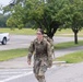 A female soldier in full military camouflage (OCP/MultiCam) uniform is walking along a paved road or parking area on what appears to be a military installation. She is carrying a large rucksack on her back and wearing a plate carrier or chest rig, suggesting she may be participating in a ruck march or physical training event. A white pickup truck is visible in the background along with manicured grass and trees.