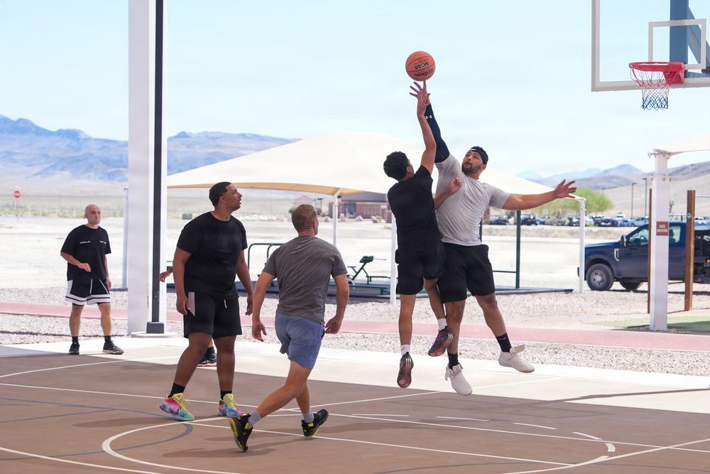A group of men are playing an outdoor basketball game on a paved court in what appears to be a desert environment, likely in the American Southwest based on the arid landscape and mountains in the background. Two players are jumping and contesting for the ball near the basket, while three other players are positioned nearby. The facility appears modern and well-maintained, with shade structures and a parking area visible in the background.