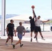 A group of men are playing an outdoor basketball game on a paved court in what appears to be a desert environment, likely in the American Southwest based on the arid landscape and mountains in the background. Two players are jumping and contesting for the ball near the basket, while three other players are positioned nearby. The facility appears modern and well-maintained, with shade structures and a parking area visible in the background.