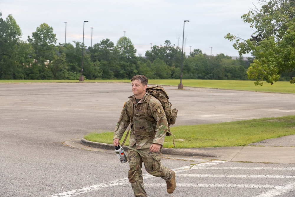 A soldier in US Army Operational Camouflage Pattern (OCP) uniform is running across a parking lot while carrying a large rucksack on his back and a water bottle in his hand. The setting appears to be a military installation or base, identifiable by the large open parking area, light poles, and maintained grounds. The activity appears to be a ruck march or physical fitness training exercise.