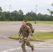 A soldier in US Army Operational Camouflage Pattern (OCP) uniform is running across a parking lot while carrying a large rucksack on his back and a water bottle in his hand. The setting appears to be a military installation or base, identifiable by the large open parking area, light poles, and maintained grounds. The activity appears to be a ruck march or physical fitness training exercise.