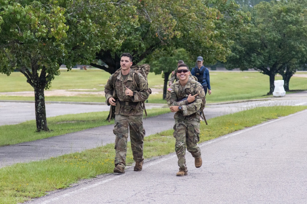 Two U.S. Army soldiers in MultiCam (OCP) camouflage uniforms are conducting a ruck march along a paved road on what appears to be a military installation. Both soldiers are carrying large rucksacks and appear to be in good spirits, with the soldier on the right smiling broadly. Additional personnel are visible in the background on the same road.